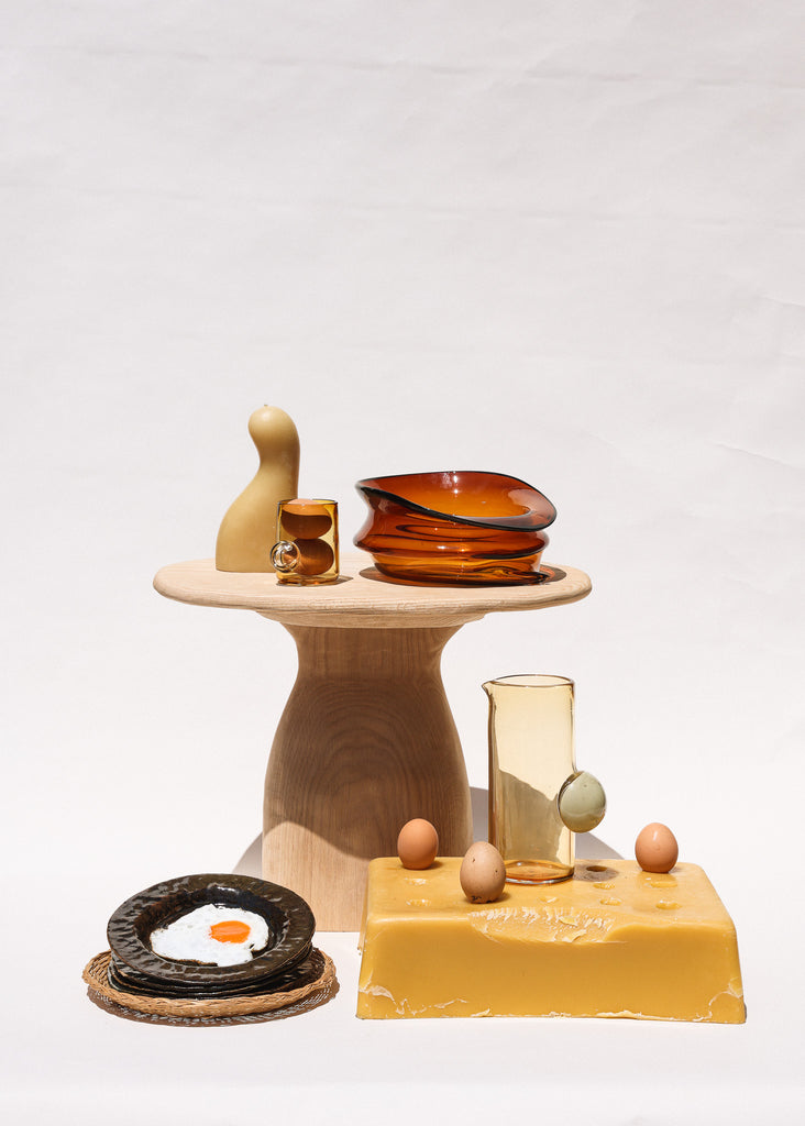 Curated tableware items including an amber glass, an orange bowl, wooden stand, clear pitcher with a green handle, fried egg on a dark plate, and eggs on a yellow block.